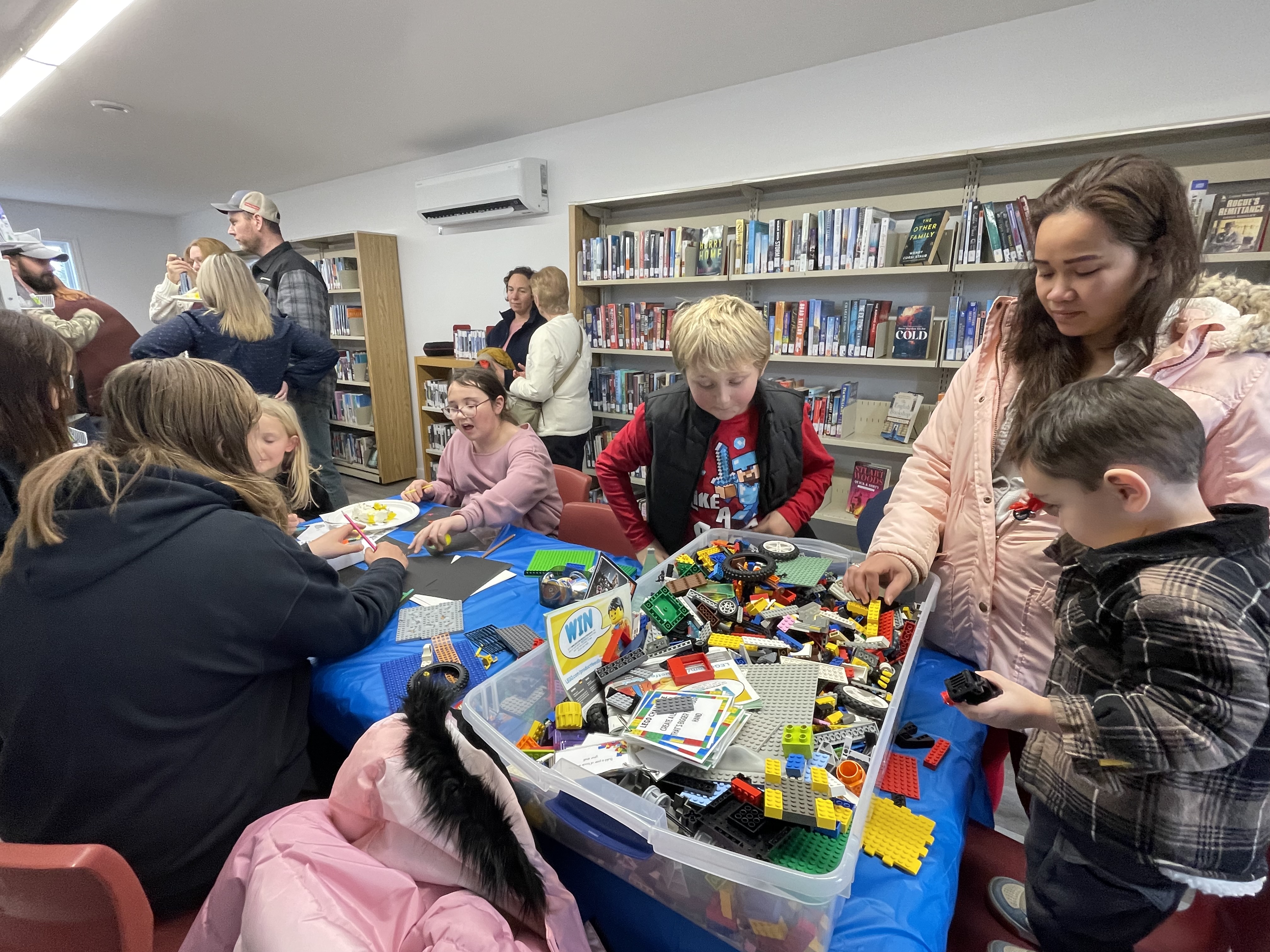 A group of children crowd around a table that has bins of Lego building blocks.